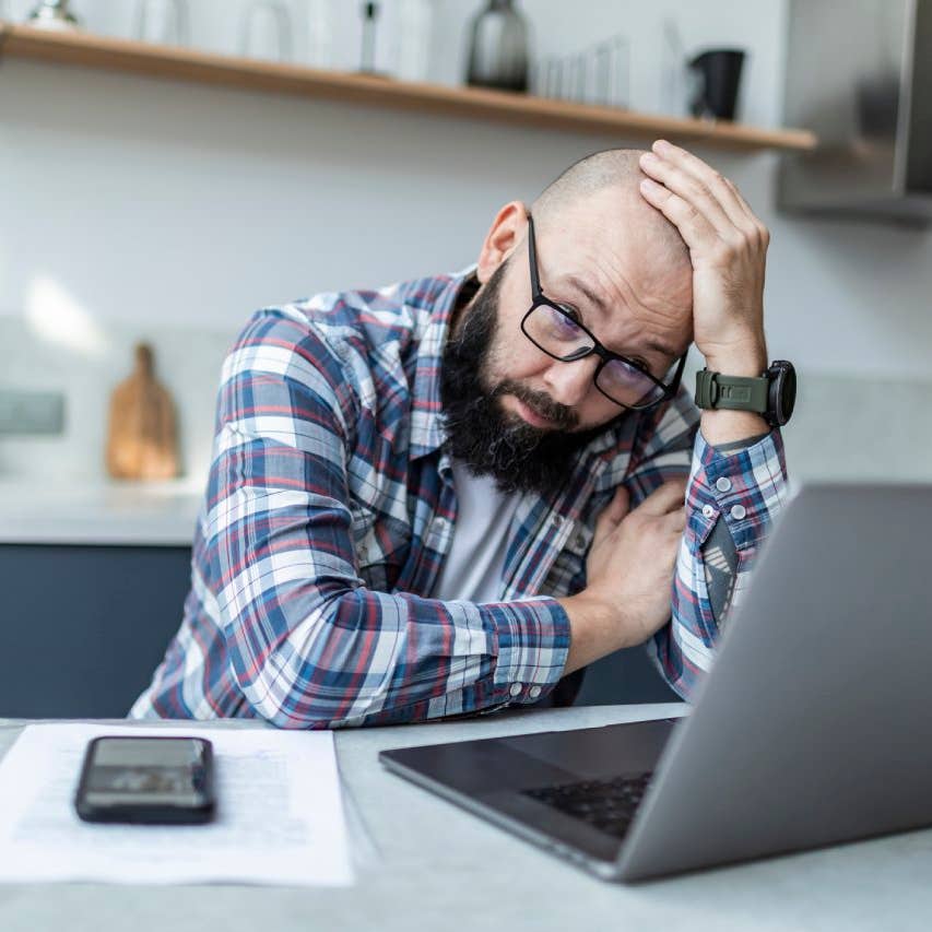 Adult man sitting at the kitchen with laptop and looking upset with the news.