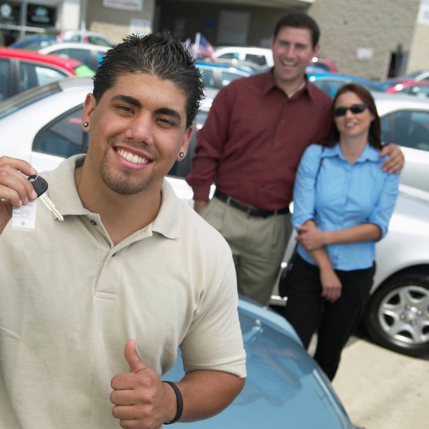 Young man at car lot, holding keys, parents in background, portrait