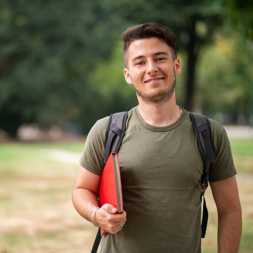 Smiling university student walking in park with backpack and book